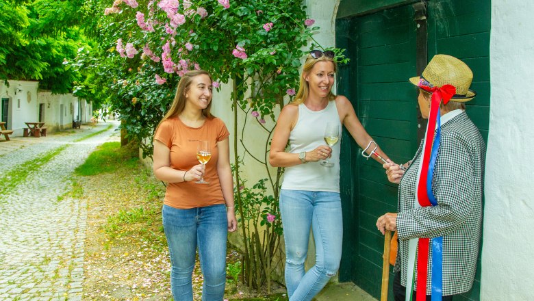 Vino Versum Poysdorf, © Semrad Drei Personen stehen vor einem grünen Tor, zwei halten Weingläser, eine trägt einen Strohhut mit bunten Bändern.