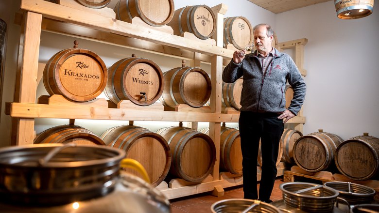 Kraxados und Whisky, © Werner Kölbel Ein Mann steht in einem Raum mit Holzfässern, die mit 'Kraxados' und 'Whisky' beschriftet sind, und hält ein Glas in der Hand.