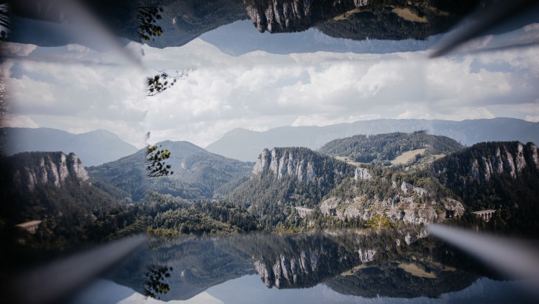 Semmeringbahnwanderweg, © Wiener Alpen/nicoleseiser.at Spiegelbild einer Berglandschaft mit Viadukt und bewaldeten Hügeln unter blauem Himmel.