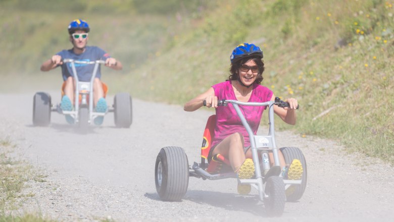 Mönichkirchen, © Erlebnisalm Mönichkirchen/Fotograf & Fee Zwei Personen fahren auf Dreirädern einen staubigen Weg hinunter.
