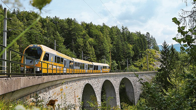 Mariazellerbahn, © weinfranz.at Ein gelber Zug der Mariazellerbahn fährt über eine Steinbrücke in einer bewaldeten Landschaft.