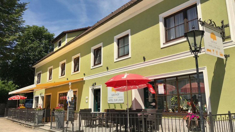 Gasthof zur Linde, © Matthias Schön Ein grüner Gasthof mit Terrasse und roten Sonnenschirmen, beschriftet mit 'Gasthof zur Linde'.