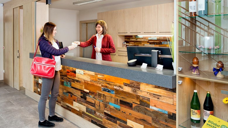 Check-In Bereich, © foto.hoefinger.at Zwei Frauen am Check-In-Schalter, eine überreicht der anderen ein Dokument. Der Schalter ist aus Holz, daneben ein Regal mit Getränken und Dekoration.
