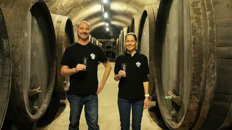 Historischer Fasskeller der Sektkellerei, © Sektkellerei Inführ Zwei Personen stehen in einem Weinkeller mit großen Holzfässern und halten Sektgläser.