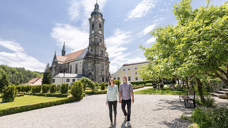 Stift Zwettl, © Stift Zwettl/Studio Kerschbaum Ein Paar spaziert im Garten des Stifts Zwettl, im Hintergrund die Kirche und ein blauer Himmel.