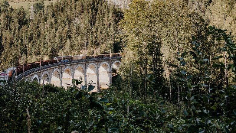 Semmeringbahnwanderweg, © Wiener Alpen/nicoleseiser.at Ein Zug fährt über ein Viadukt in einer bergigen, bewaldeten Landschaft unter blauem Himmel.