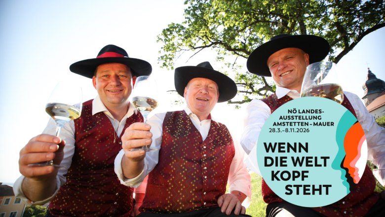 Mostbarone, © Mostviertel Tourismus/schwarz-koenig.at Drei Männer in traditioneller Kleidung mit Weingläsern, lächelnd unter einem Baum. Logo der NÖ Landesausstellung 2026 rechts unten.