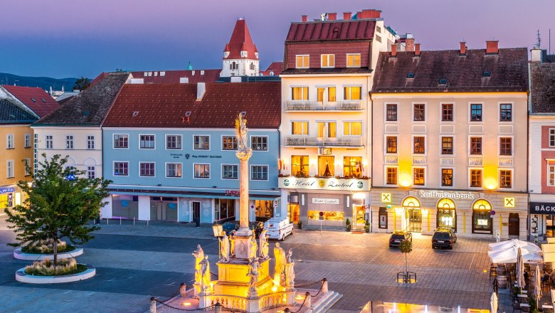 Hotel Zentral, © Martin Haider Fotografie Abendliche Stadtansicht mit beleuchtetem Brunnen und Gebäuden, darunter ein Hotel und eine Bank.