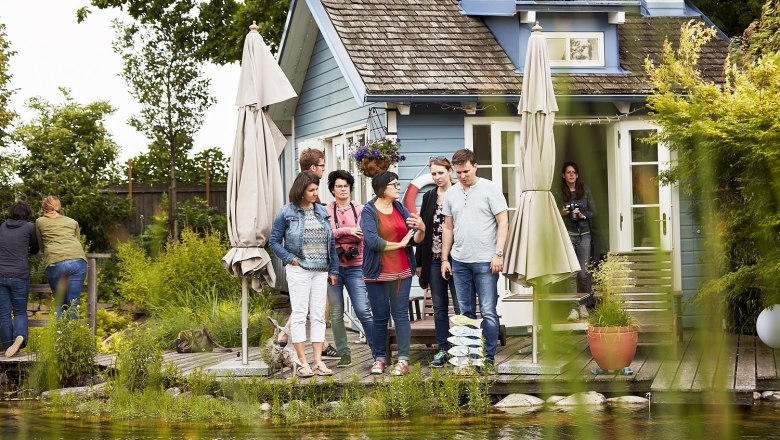 Gartenführung, © Oskar Hemm Eine Gruppe von Menschen steht vor einem kleinen blauen Haus mit Terrasse und Sonnenschirmen, umgeben von Pflanzen.