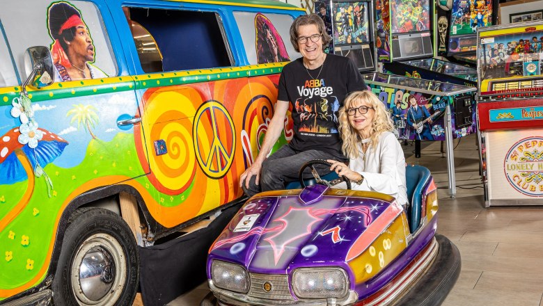 Ein bunt bemalter VW-Bus mit Peace-Zeichen und ein lila Autoskooter. Zwei Personen posieren fröhlich davor. Im Hintergrund sind Flipperautomaten zu sehen.