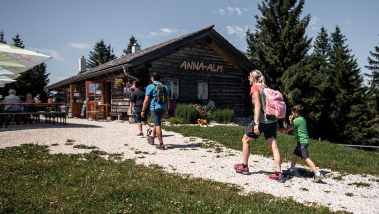 Wandernde Familien und Ausflugsg&auml;ste erreichen die gem&uuml;tliche Anna-Alm inmitten der Berglandschaft &ndash; ein beliebter Einkehrort mit uriger H&uuml;ttenatmosph&auml;re.