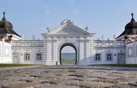 Barockes Tor mit weißen Verzierungen und Figuren, gepflasterter Weg im Vordergrund.