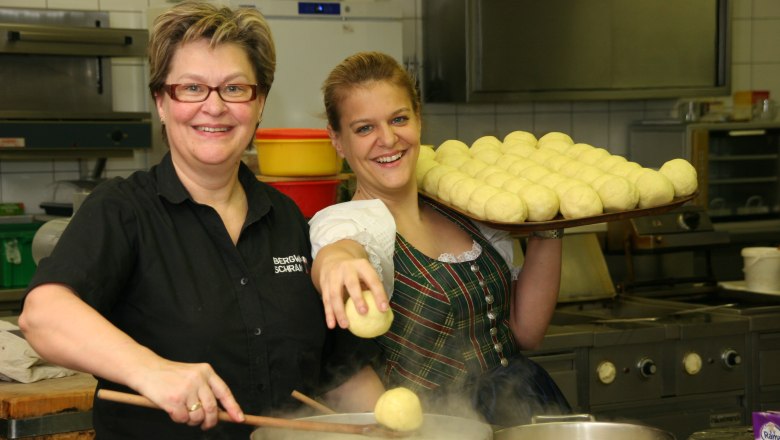 Zwei Frauen in einer Küche, eine hält einen Tablett mit Knödeln, die andere kocht Knödel in einem Topf.