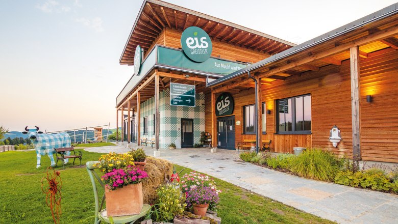 Gebäude von Eis Greissler mit Holzfassade und dekorativer Kuhfigur im Garten.