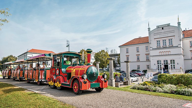Ein bunter Touristenzug f&auml;hrt an einem wei&szlig;en Geb&auml;ude mit der Aufschrift 'Rathaus' vorbei.
