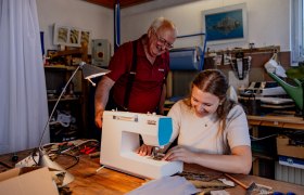 Eine junge Frau näht an einer Nähmaschine, während ein älterer Mann lächelnd zuschaut.