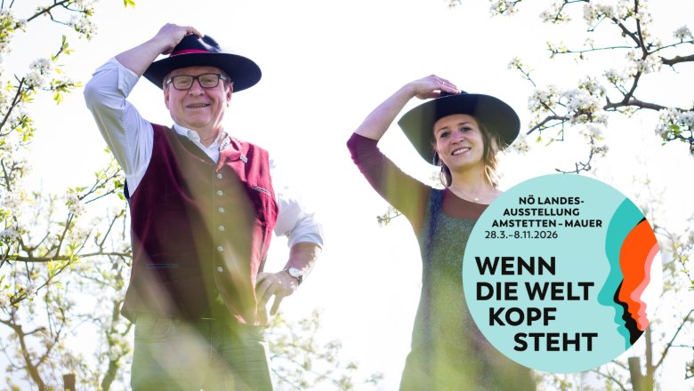 Zwei Personen mit H&uuml;ten in einem bl&uuml;henden Garten, Plakat f&uuml;r N&Ouml; Landesausstellung 2026.