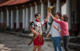 Ein Mann in römischer Rüstung hält einen Helm, während zwei Personen ihn bewundern.