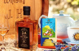 Eine Flasche Waldviertler Rye Whisky neben einem Glas, Teepackung und Teekanne auf einem Tisch mit Lavendel und Getreide.