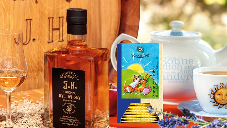 Eine Flasche Waldviertler Rye Whisky neben einem Glas, Teepackung und Teekanne auf einem Tisch mit Lavendel und Getreide.