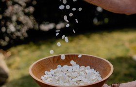 Eine Hand h&auml;lt eine Holzschale mit wei&szlig;en Kn&ouml;pfen, w&auml;hrend weitere Kn&ouml;pfe von oben hineinfallen.
