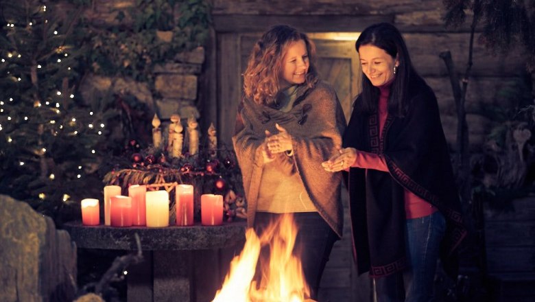 Zwei Frauen stehen lächelnd an einem Feuer, umgeben von Kerzen und Weihnachtsdekoration.