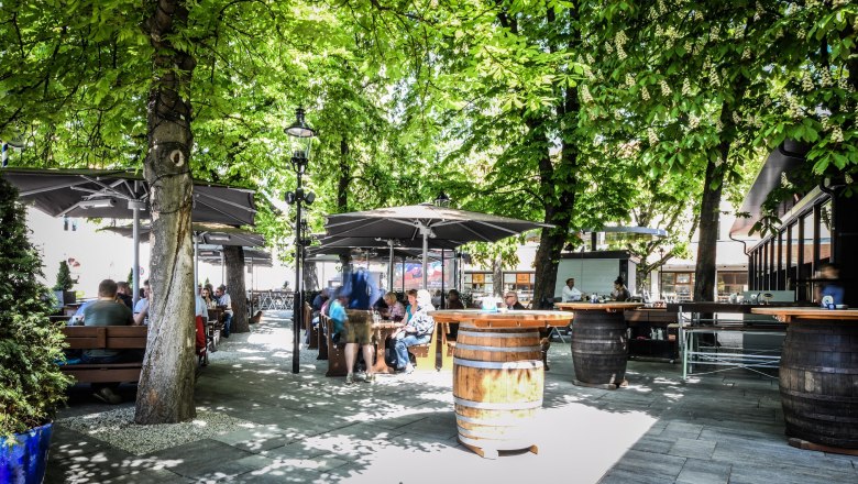 Ein Biergarten mit Holztischen und -fässern unter grünen Bäumen, Menschen sitzen und genießen die Atmosphäre.