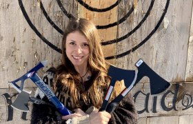 Eine Frau h&auml;lt drei &Auml;xte vor einer Holzscheibe mit Zielscheibe.