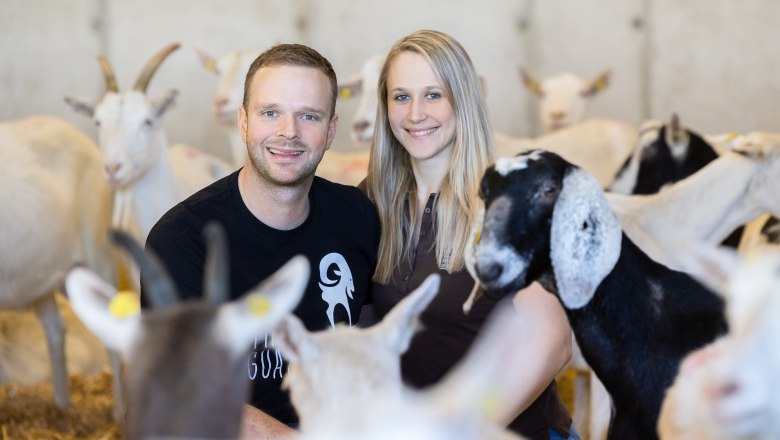 Ein Mann und eine Frau lächeln inmitten von Ziegen in einem Stall.