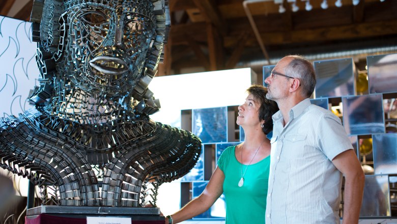 Zwei Personen betrachten eine große Metallskulptur in einem Ausstellungsraum.