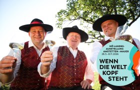 Drei M&auml;nner in traditioneller Kleidung mit Weingl&auml;sern, l&auml;chelnd unter einem Baum. Logo der N&Ouml; Landesausstellung 2026 rechts unten.