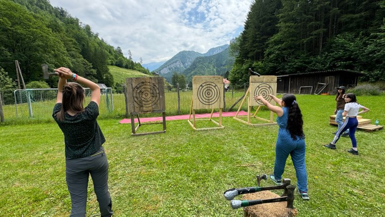 Personen werfen Äxte auf Zielscheiben in einer grünen, bergigen Landschaft.