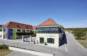 Geb&auml;ude der Winzer Krems mit rotem Dach und gr&uuml;nen Fensterl&auml;den, umgeben von Weinbergen.