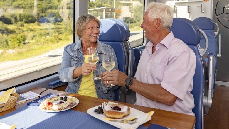 Ein älteres Paar stößt mit Weingläsern in einem Zug an, mit Essen auf dem Tisch.
