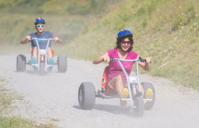 Zwei Personen fahren auf Dreirädern einen staubigen Weg hinunter.