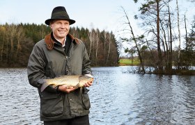 Mann mit Hut hält einen Karpfen vor einem Teich.