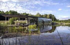 Modernes Geb&auml;ude mit Glasfassade am Teich, umgeben von Natur.