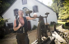 Ein Mann in traditioneller Kleidung steht vor einem historischen Geb&auml;ude mit Werkzeugen.