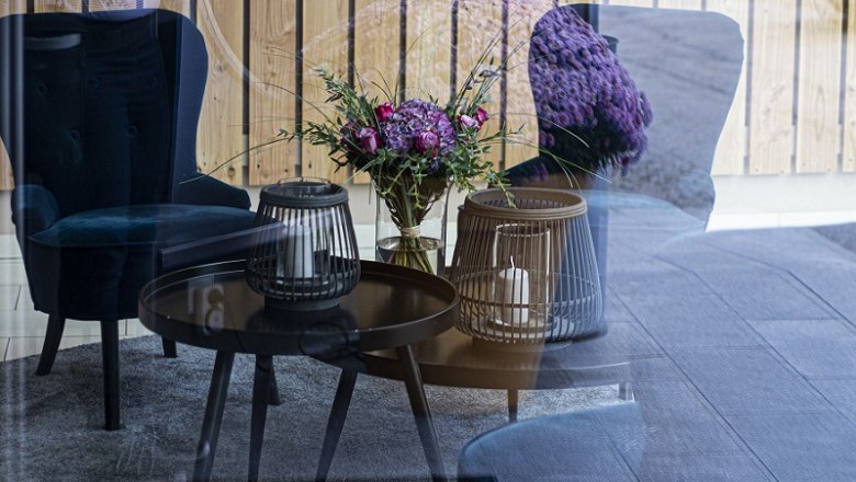 Ein gem&uuml;tlicher Loungebereich mit blauen Sesseln, einem Tisch mit Kerzen und einem Blumenstrau&szlig;, reflektiert in einer Glaswand.