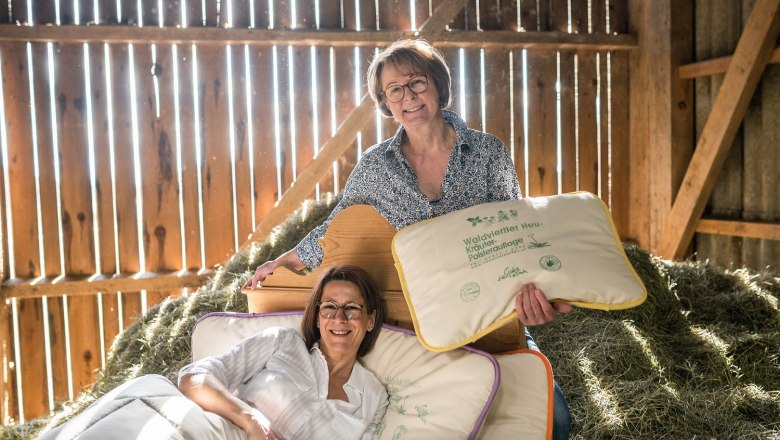 Zwei Frauen in einer Scheune mit Heu, eine liegt auf einem Bett, die andere hält ein Kissen.