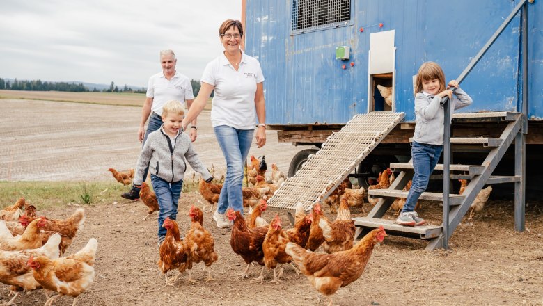 Familie mit H&uuml;hnern vor einem blauen H&uuml;hnerstall auf einem Feld.