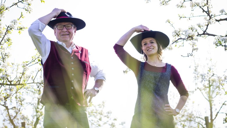 Zwei Personen in traditioneller Kleidung mit Hüten stehen in einem blühenden Obstgarten.