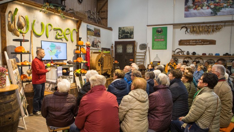 Ein Mann hält einen Vortrag vor einer Gruppe von Menschen in einem rustikalen Raum mit Kürbissen und landwirtschaftlichen Werkzeugen.