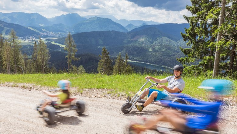 Personen fahren auf Dreir&auml;dern einen H&uuml;gel hinunter mit Berglandschaft im Hintergrund.
