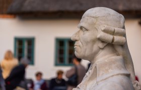 B&uuml;ste eines Mannes im Profil vor einem Geb&auml;ude mit Menschen im Hintergrund.