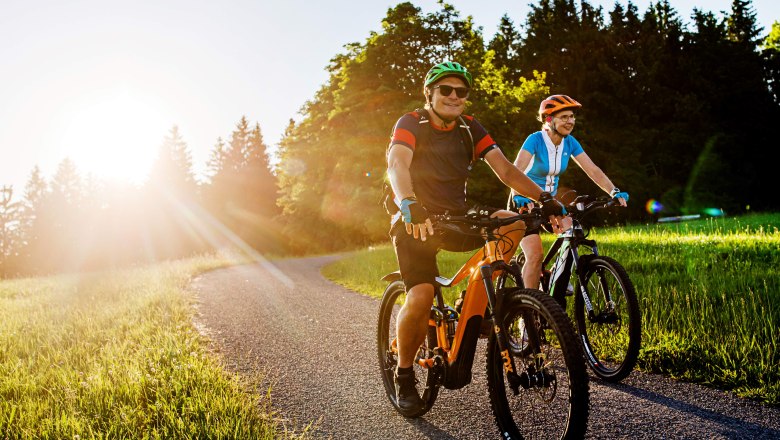 Zwei Personen fahren bei Sonnenuntergang auf E-Bikes auf einem Weg durch eine gr&uuml;ne Landschaft.