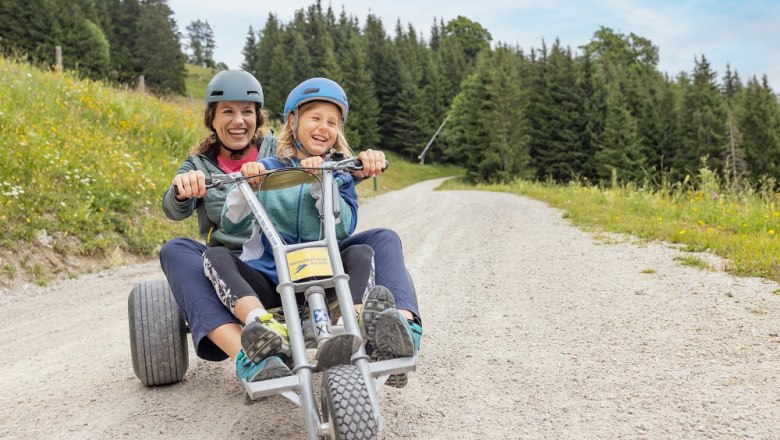 Zwei Personen fahren auf einem Mountaincart auf einem Schotterweg, umgeben von Wiesen und Wald.