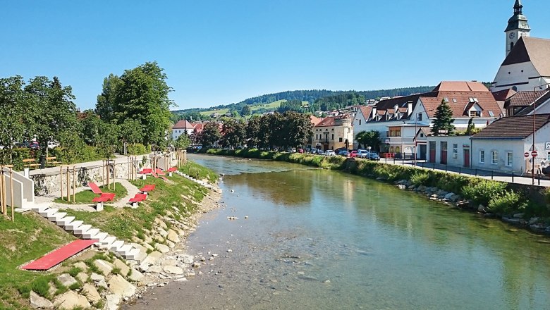 Fluss mit Uferpromenade und roten Liegen, Stadt im Hintergrund.
