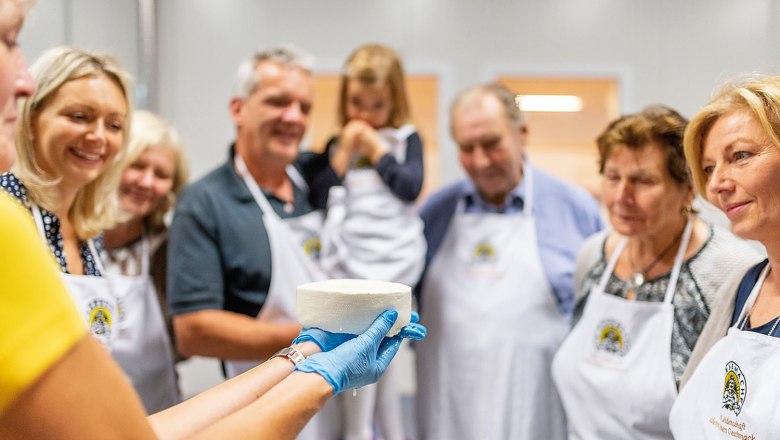 Eine Gruppe von Menschen betrachtet einen frisch hergestellten Käse, der von einer Person mit blauen Handschuhen gehalten wird.